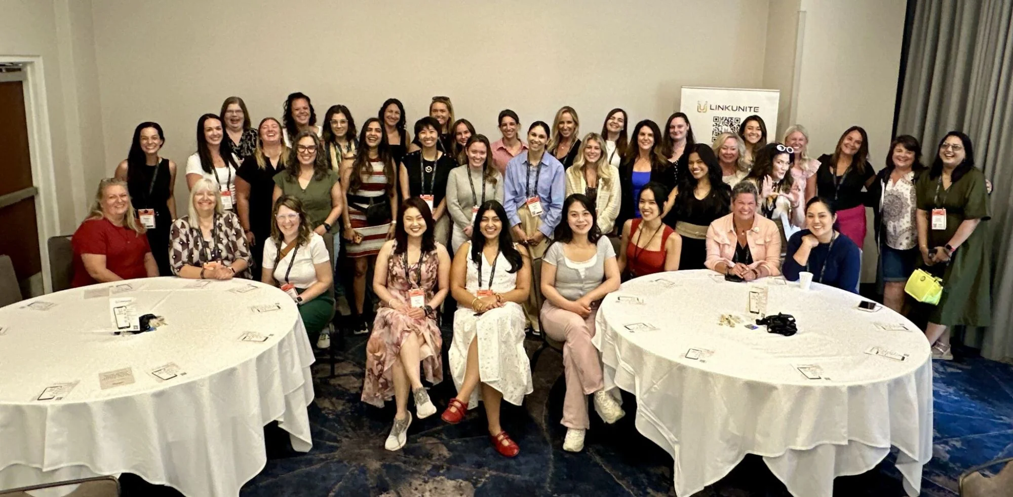 A full room of attendees smiling for a group photo at the LinkUnite L.I.N.K & Learn session, seated around two round tables.